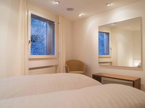 Ein Schlafzimmer mit Bett, Stuhl, Nachttisch, Fenster mit Vorhängen und Spiegel an der Wand.
