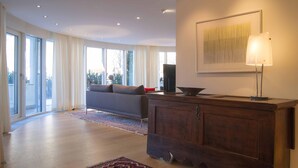 A spacious living room with a sofa, a wooden cabinet, and a large window. - Residenza Alpenrose 31 – Spacious 4.5-room flat with a terrace and mountain view (Sils / Segl Maria)