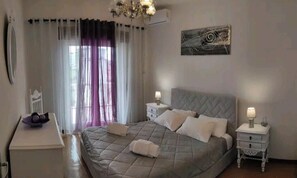 A bedroom with a bed, two bedside tables, a nightstand, a chandelier, a window with curtains, and a wall art piece.