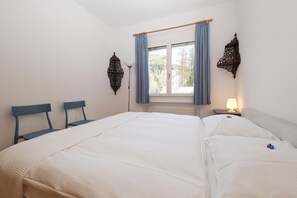 Ein Schlafzimmer mit einem Bett, zwei Stühlen, einem Fenster mit Vorhängen, einer Wandleuchte und einem Nachttisch mit Lampe.