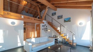 Ein geräumiger Wohnbereich mit hohen Decken, Holzbalken, einer Treppe, einem Sofa, einem Couchtisch und Blick ins Freie.