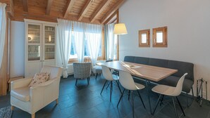 Ein gemütliches Zimmer mit Holzbalkendecke, einem Esstisch mit Stühlen, einer Couch und einem Fenster mit Vorhängen.