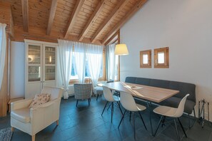 Ein gemütliches Zimmer mit Holzbalkendecke, einem Esstisch mit Stühlen, einer Couch und einem Fenster mit Vorhängen.