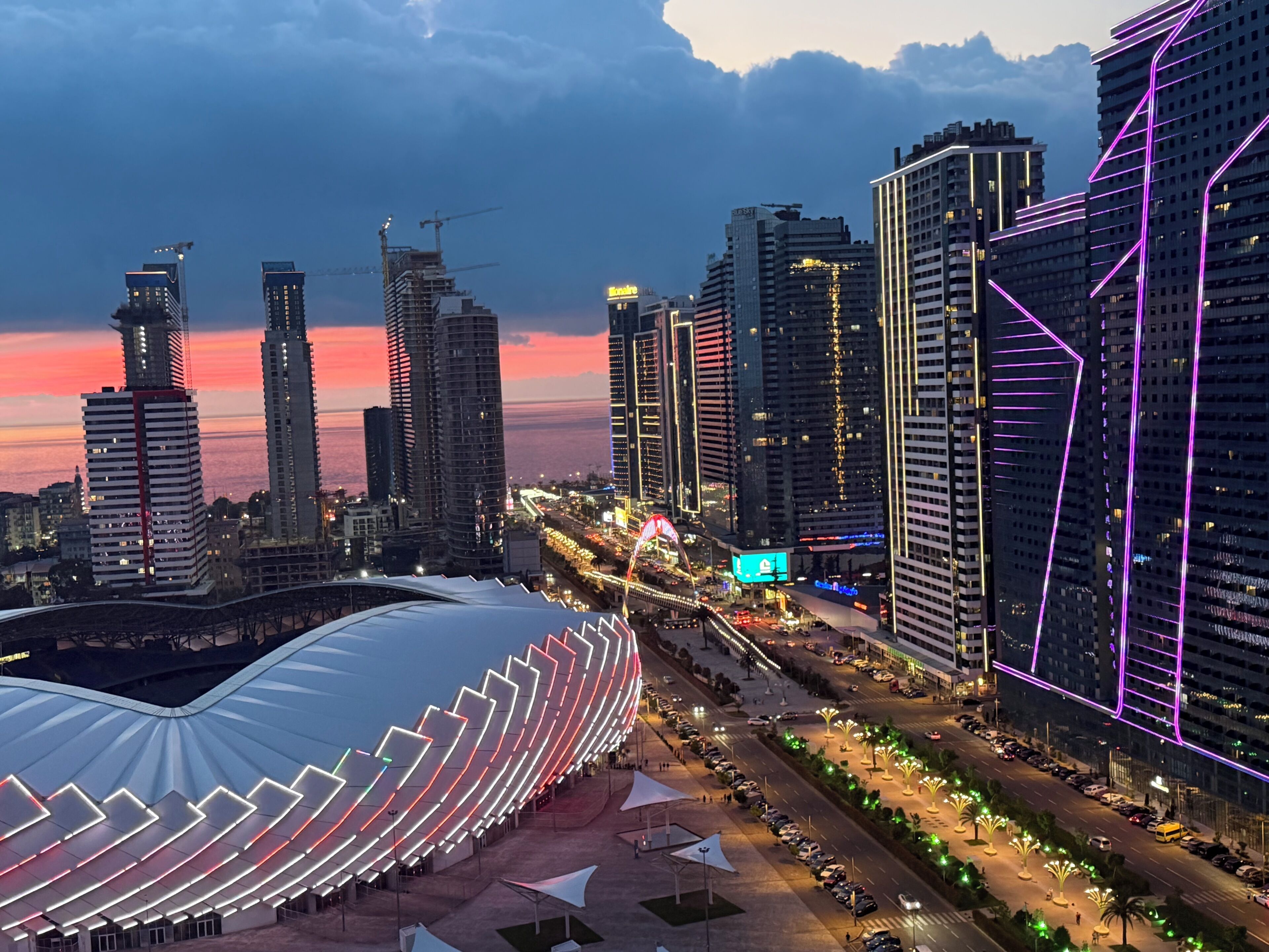 A modern cityscape with high-rise buildings, a stadium, and a well-lit road.