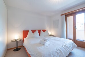 A hotel room with a bed, a bedside table, a lamp, and a window. - Chesa Polaschin – Bright Apartment with a Balcony & Prime Location (Sils/Segl Maria)