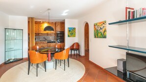 Une cuisine moderne avec des meubles en bois, un coin repas avec des chaises orange, un meuble vitré et un téléviseur posé sur un meuble.