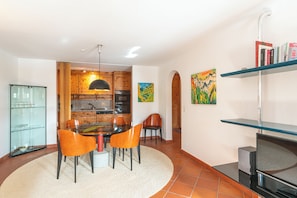 A modern kitchen with wooden cabinets, a dining area with orange chairs, a glass cabinet, and a TV on a stand. - Chesa Polaschin – Bright Apartment with a Balcony & Prime Location (Sils/Segl Maria)