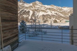 Ein mit Schnee bedeckter Balkon mit Blick auf die Berge, Holzhüttenwände und ein Metallgeländer.