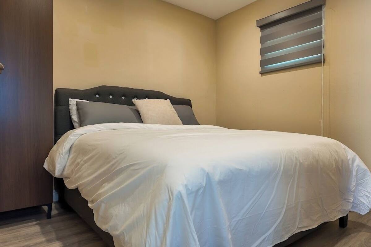 A bedroom with a bed, a headboard, a window with blinds, and a wooden floor.