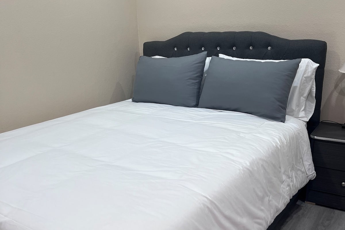A neatly made bed with white bedding and two pillows, a dark headboard, a bedside table with a lamp, and a wooden floor.