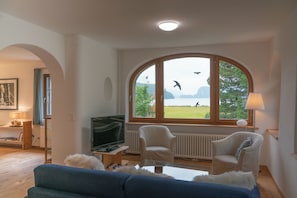 Ein Wohnzimmer mit einem großen Bogenfenster, einem Fernseher, zwei weißen Ohrensesseln, einem blauen Sofa und einer Lampe.