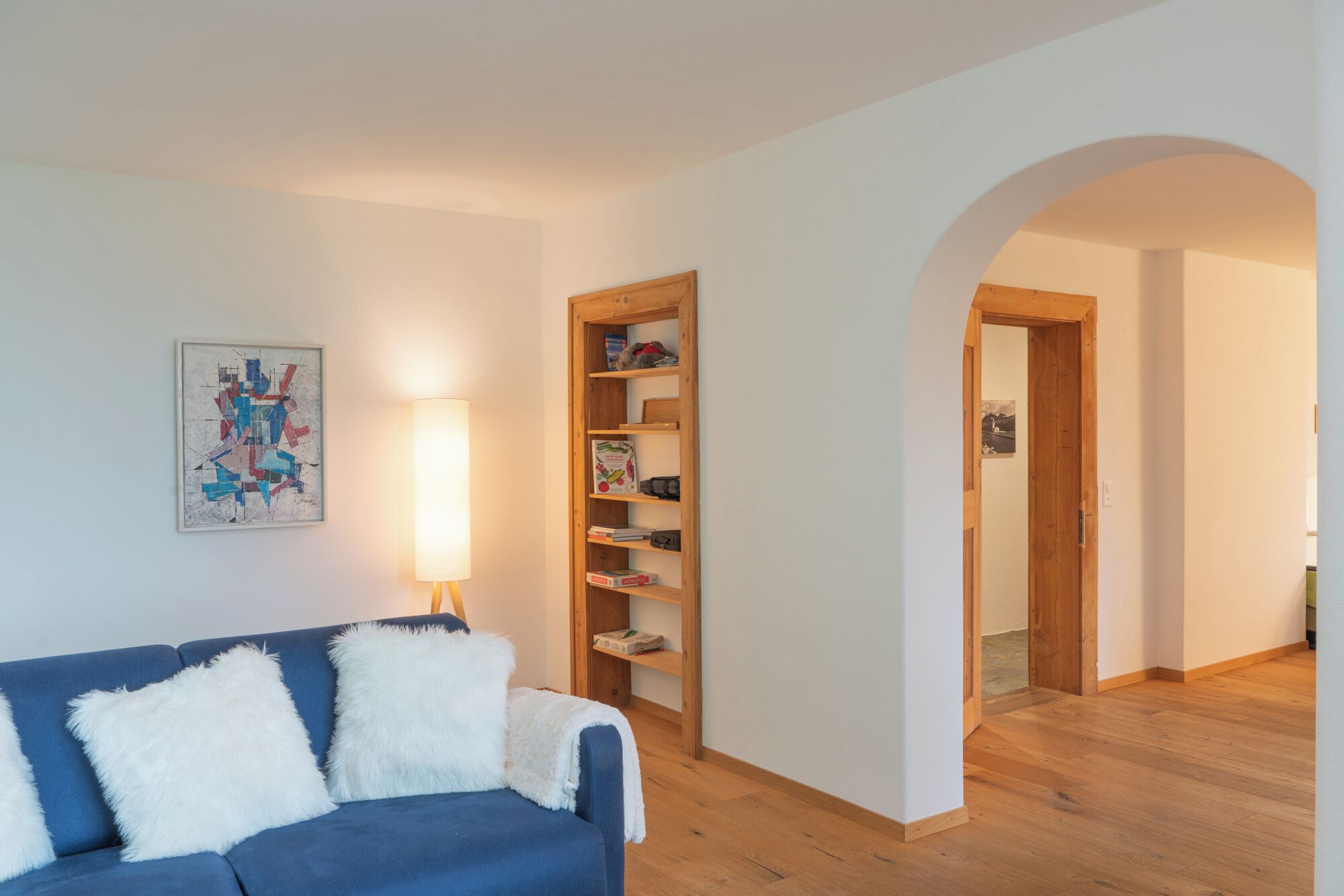 Una sala de estar con un sofá azul, un almohadón blanco y mullido, una lámpara, una estantería y un arco que da a otra habitación.