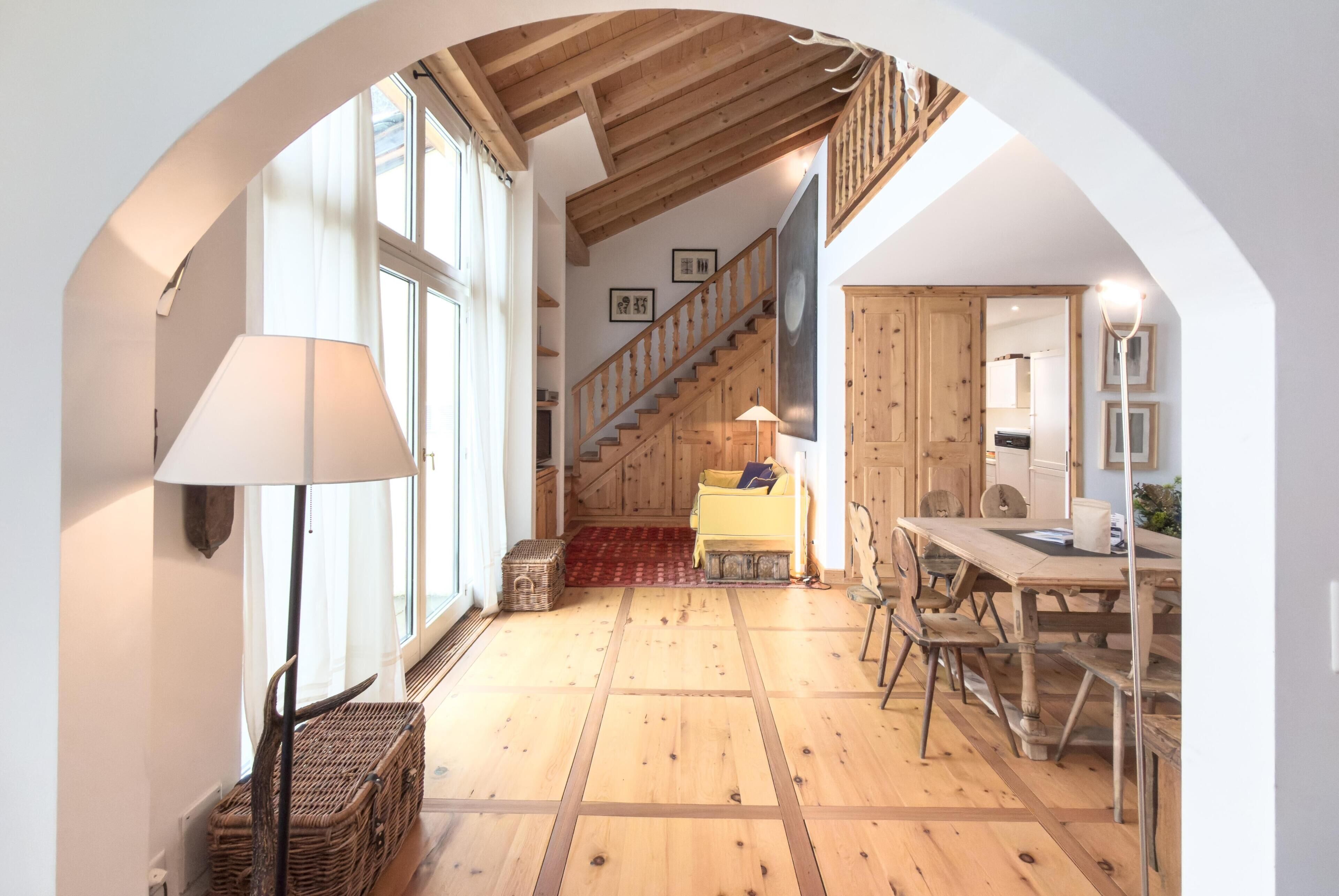 Una habitación amplia con piso de madera, una escalera, un comedor y un ventanal grande.