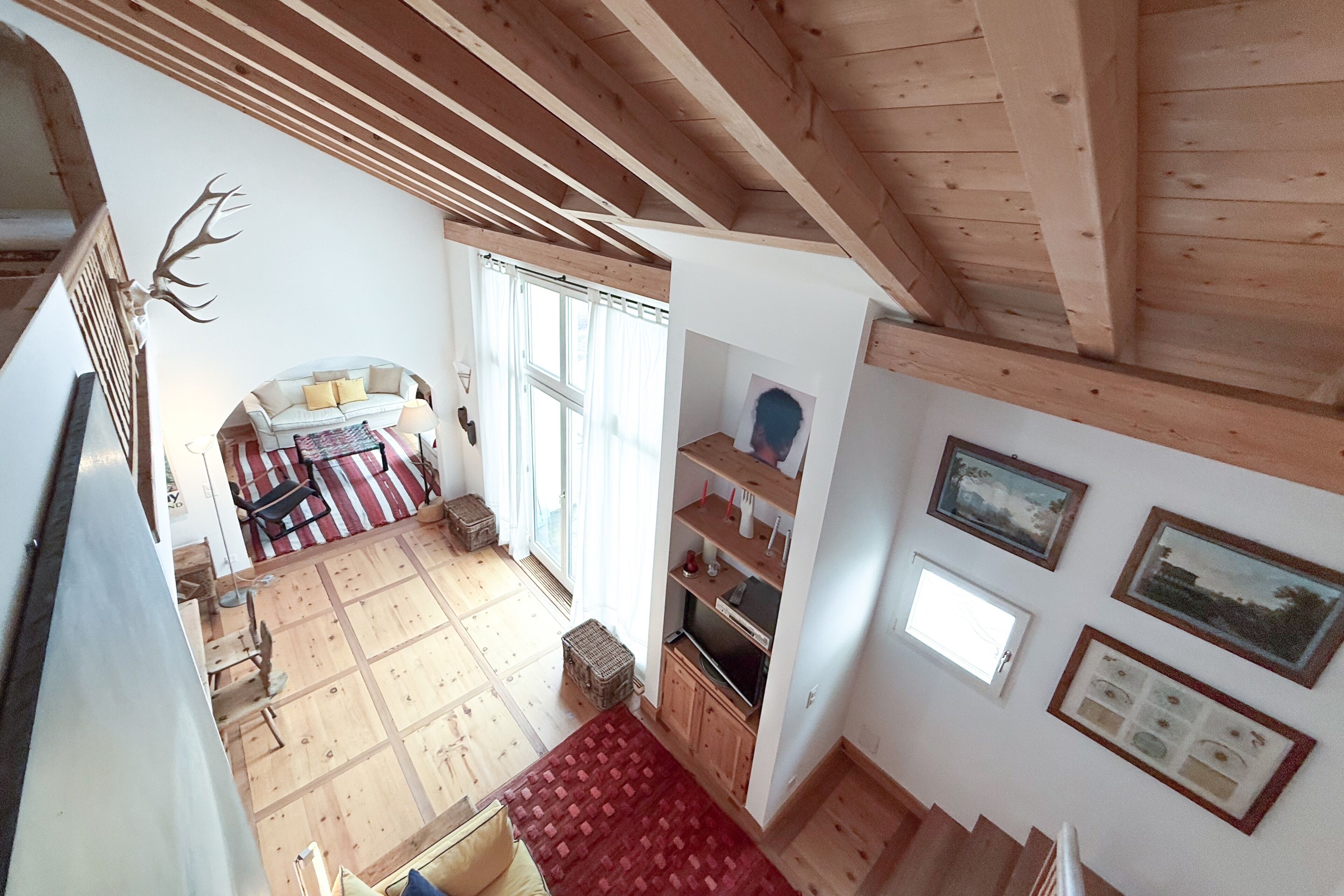A spacious room with a high ceiling, wooden beams, a sofa with cushions, a television, and a red rug.