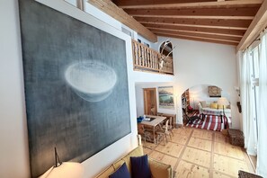 Ein geräumiges Zimmer mit einem großen abstrakten Gemälde, einer Holzbalkendecke, einem Balkon mit einer Hirschskulptur, einem Essbereich und einer gemütlichen Sitzecke mit einem Streifen Teppich.