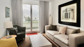A modern living room with a sofa, armchair, and coffee table, a large window with curtains, and a framed picture on the wall. - Residenza Alpenrose 19 – Stylish flat with 2 terraces & panoramic views (Sils/Segl Maria)