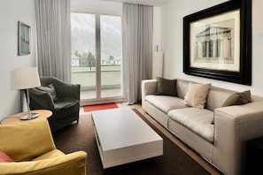 Ein modernes Wohnzimmer mit einer Couch, einem Sessel und einem Couchtisch, einem großen Fenster mit Vorhängen und einem gerahmten Bild an der Wand.
