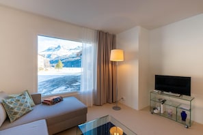 Ein modernes Hotelzimmer mit einer Couch, einem Glastisch, einem Fernseher auf einer Glasschrank und Blick auf schneebedeckte Berge.