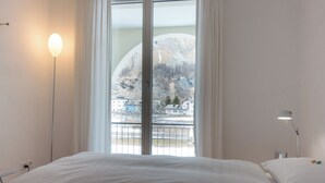 Un dormitorio con una cama, una lámpara de noche, una ventana con vistas a una montaña y edificios.