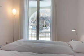 Ein Schlafzimmer mit Bett, Nachttischlampe und Fenster mit Blick auf Berg und Häuser.