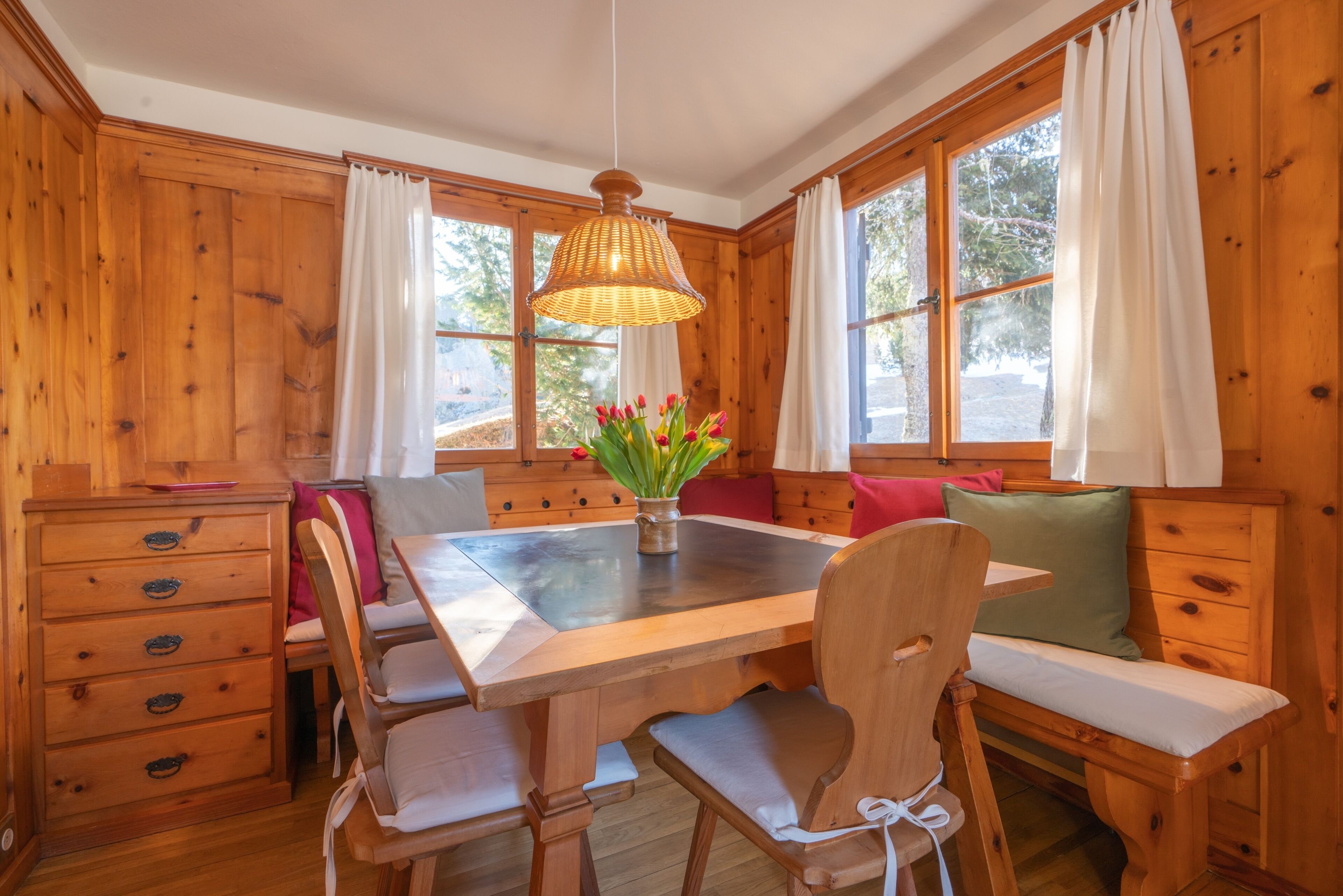 L’intérieur d’un chalet en bois comprend une table à manger, des bancs, un lustre et de grandes fenêtres.