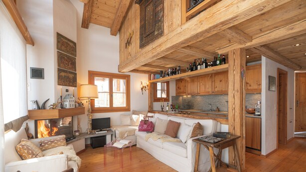 Uma sala de estar acolhedora com lareira, vigas de madeira, um sofá, uma mesa de centro e uma área de cozinha com armários de madeira e bancada.