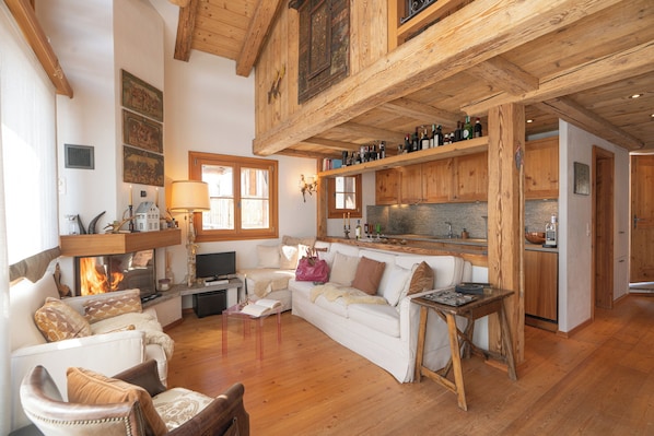 Ein gemütliches Wohnzimmer mit Kamin, Holzbalken, einer Couch, einem Couchtisch und einer Küchenzeile mit Holzschränken und einer Arbeitsplatte.