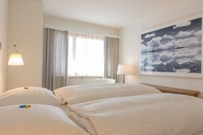 Ein Hotelzimmer mit zwei Betten, einem Fenster mit Vorhängen, einer Lampe und einem Gemälde von Wolken.