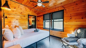 Ein Zimmer in einer Holzhütte mit einem Bett, einem Sessel, einem Deckenventilator und einem Fenster.