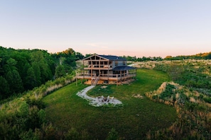A two-story wooden house with a deck, surrounded by lush greenery and a clear sky. - Nerf Blasters | Hot Tub | Mural | Arcade | Sunset (Wolfe County)