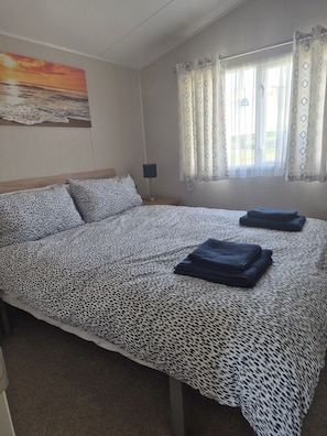 A bedroom with a bed, patterned bedding, a nightstand with a lamp, a painting on the wall, and a window with curtains. - Cabin Sleeps 4 With Garden and Parking (Eyemouth)