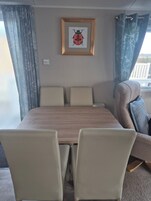 Una habitación con una mesa de madera, cuatro sillas, una obra de arte enmarcada de una mariquita y una ventana con cortinas.