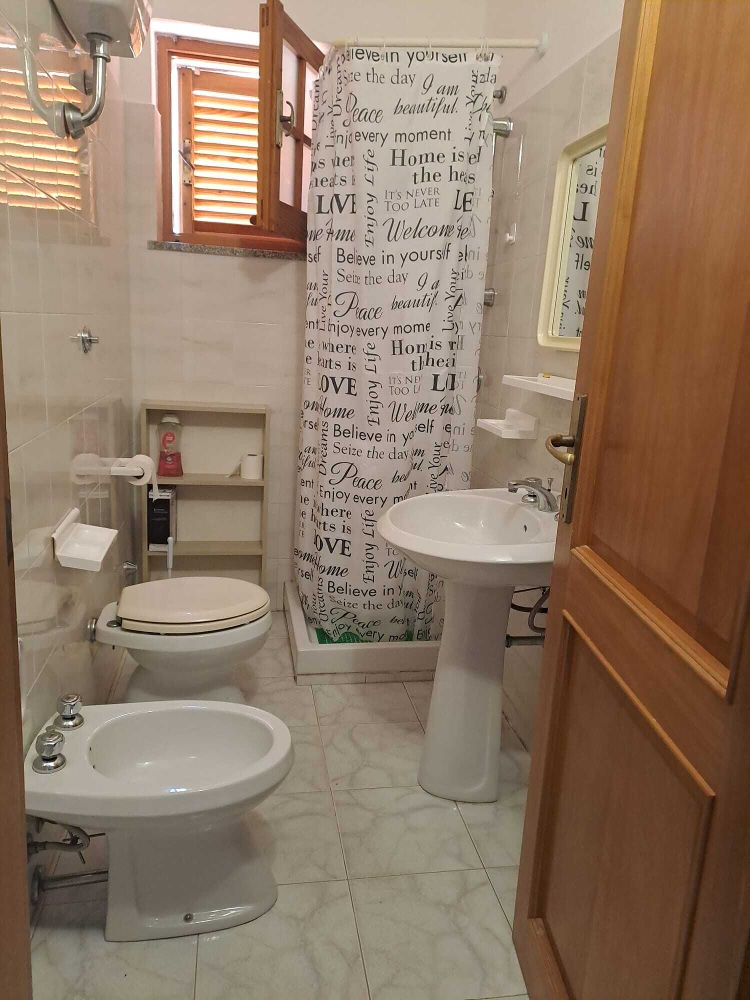 Bany amb vàter, bidé i rentapel. Cortina de dutxa amb frases motivacionals i finestra amb persianes.