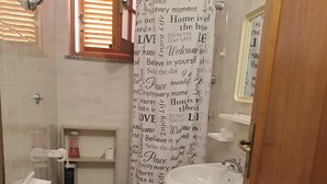 Ein Badezimmer mit Toilette, Bidet und Waschbecken, einer Duschvorhang mit motivierenden Aufschriften und einem Fenster mit Jalousien.
