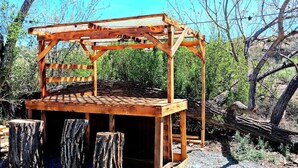 A wooden pergola structure with a slatted roof and a solid base, surrounded by natural vegetation and a fallen tree. - Journey South West Tent 3 (Farmington)