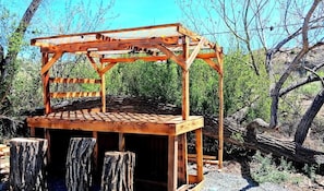 A wooden pergola structure with a slatted roof and a solid base, surrounded by natural vegetation and a fallen tree.