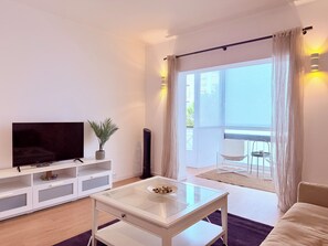 Ein modernes Wohnzimmer mit einem Flachbildfernseher, einem Glastisch, einem weißen Konsolentisch mit Pflanze und einem Balkon mit Tisch und Stühlen.
