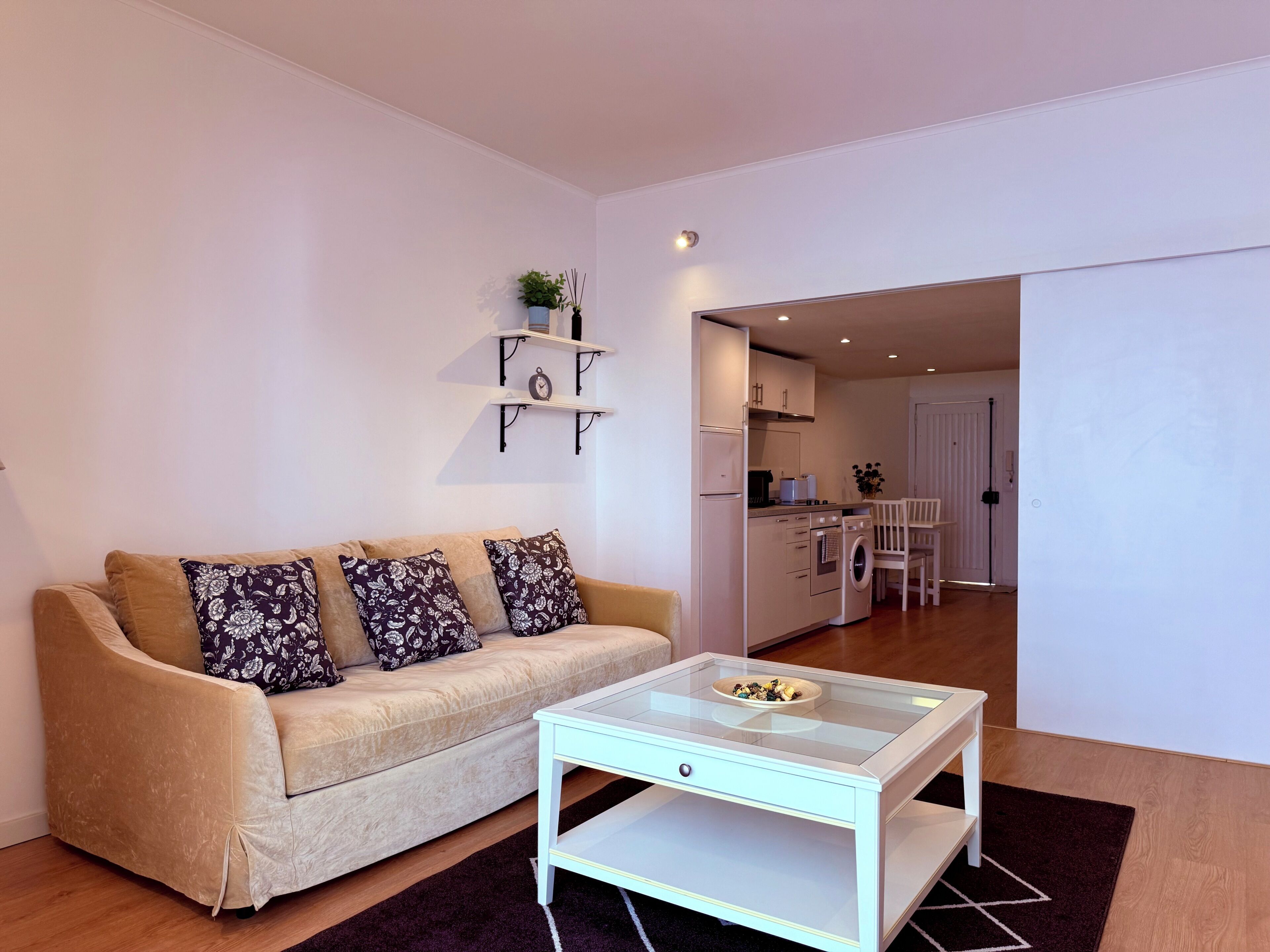 A living room with a sofa, a glass coffee table, and a kitchen in the background.