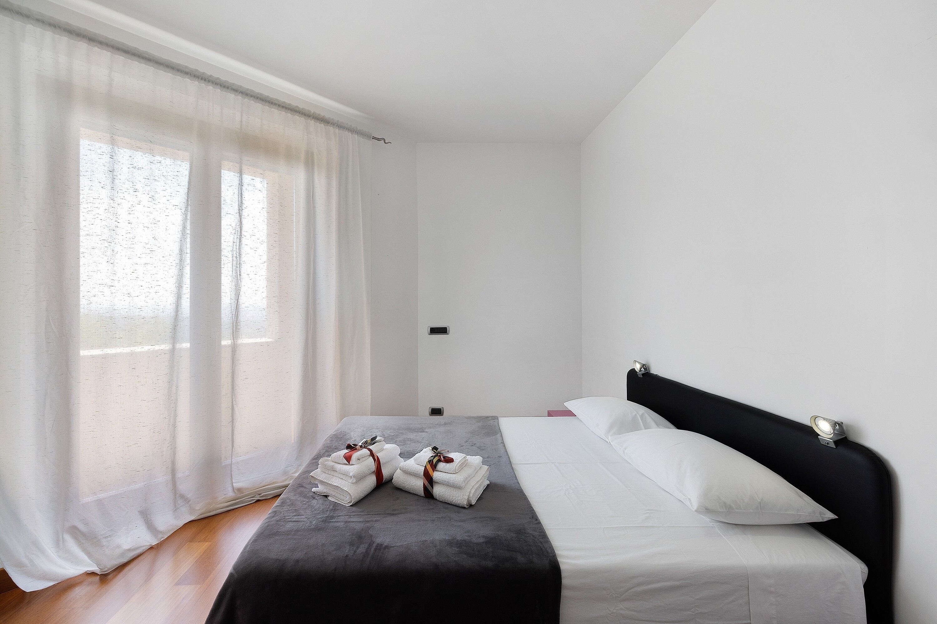 Een hotelkamer met een bed, een raam met dunne gordijnen en een nachtkastje.