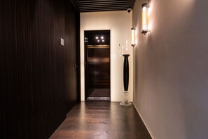 Een gang met een houten deur, een lift en een hoge staande lamp.
