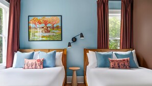 A hotel room with two beds, a small round table, a painting on the wall, and a view of greenery outside.