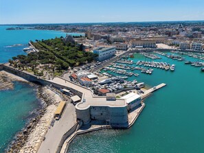 Un paisaje costero con un fuerte histórico, un puerto deportivo con numerosas embarcaciones y una zona urbana densa al fondo.