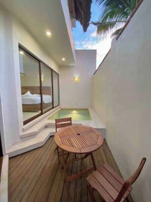 Un espacio exterior pequeño con una mesa y sillas de madera, una zona de descanso con paredes de vidrio y una piscina pequeña.