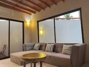 Una sala de estar moderna con un sofá, una mesa de centro redonda de madera y ventanas grandes con paneles de vidrio esmerilado.