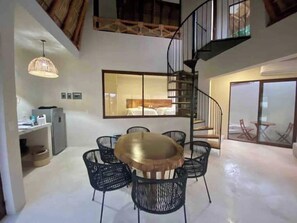 Un interno moderno con un tavolo rotondo in legno, sedie nere, una scala a chiocciola e un grande specchio che riflette una camera da letto.