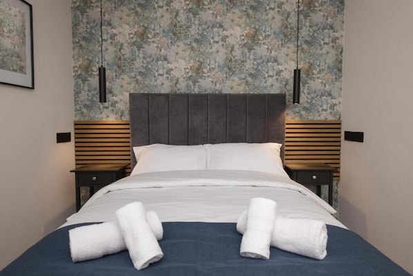 A neatly made bed with white linens and towels, a dark headboard, and a textured floral wallpaper.