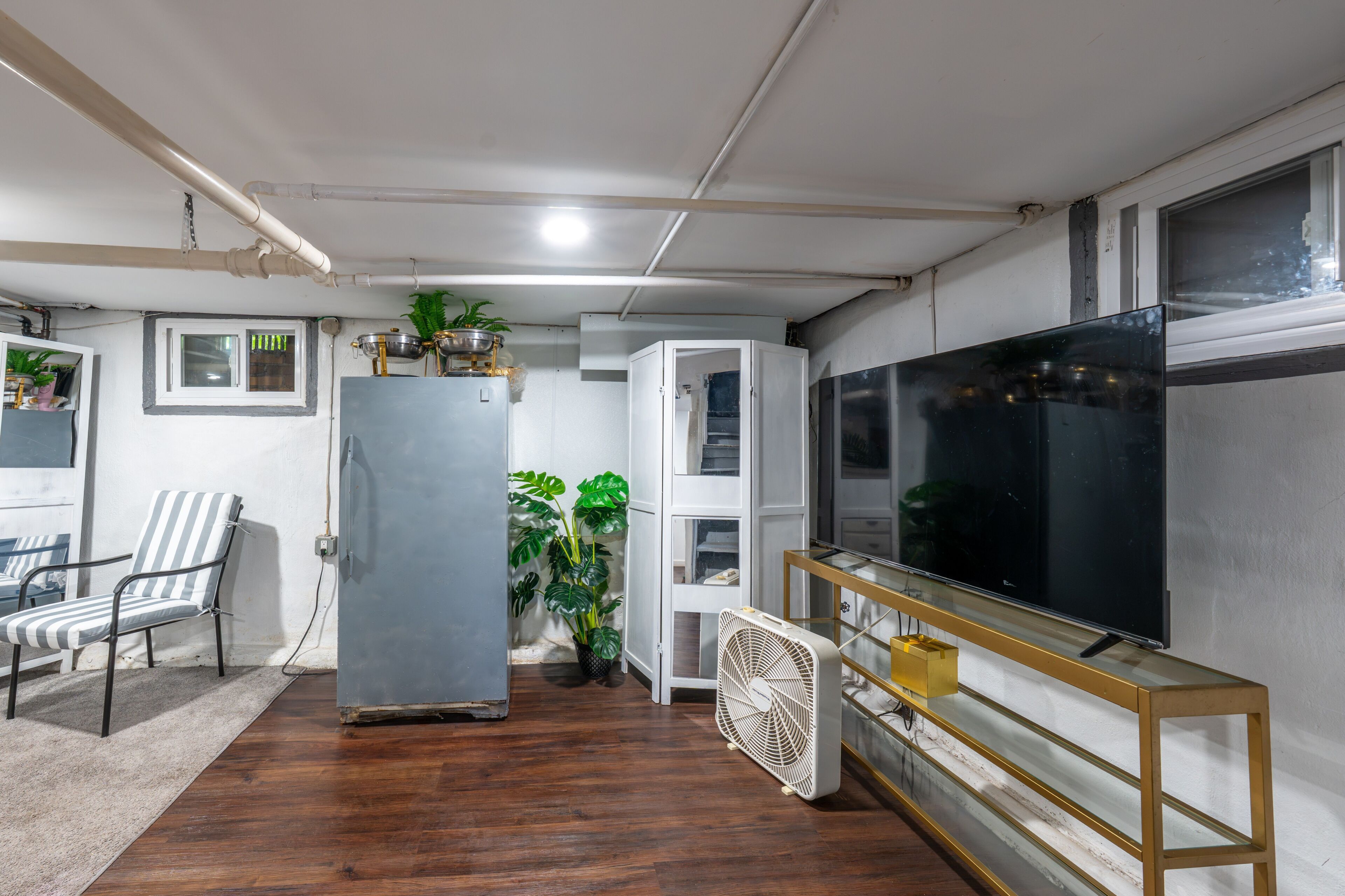 Un salon avec un grand téléviseur à écran plat, un ventilateur blanc, un fauteuil à rayures, un réfrigérateur et une plante en pot.