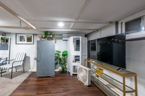 Olohuone, jossa on suuri litteänäyttöinen televisio, valkoinen tuuletin, raidallinen tuoli, jääkaappi ja ruukussa oleva kasvi.