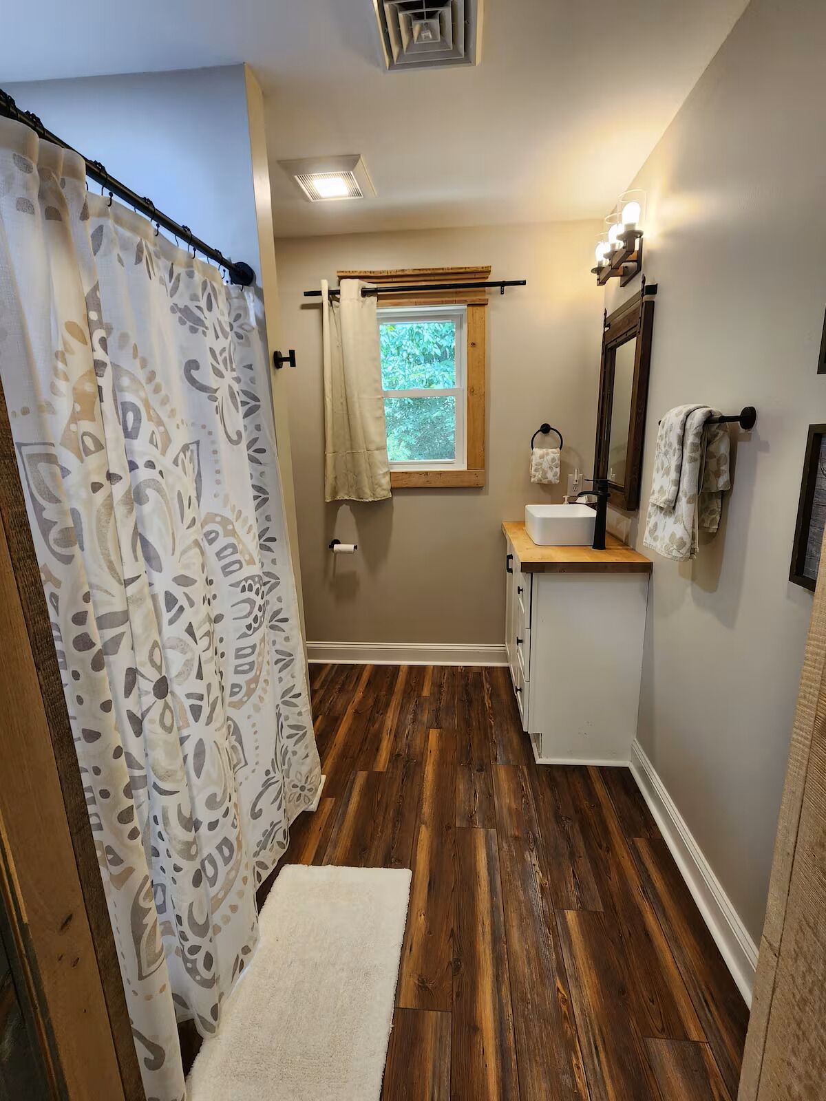 Un baño con suelo de madera, ventana, lavabo, espejo y cortina de ducha.