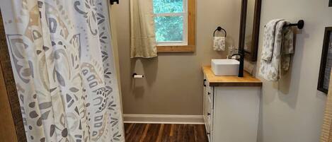 Un baño con suelo de madera, ventana, lavabo, espejo y cortina de ducha.